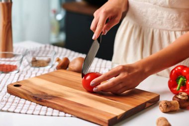 Kadın aşçı, çağdaş bir mutfakta, canlı kırmızı bir domatesi, bir dizi sebze ve aşçılık sahnesini geliştiren yemek malzemeleriyle, ahşap bir kesme tahtasıyla ustalıkla dilimliyor.