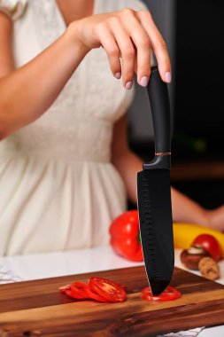 Kadın şef, renkli sebzelerle çevrili keskin siyah bir bıçakla ahşap kesme tahtasındaki taze domatesleri ustalıkla dilimliyor, canlı ve davetkar bir mutfak atmosferi yaratıyor.