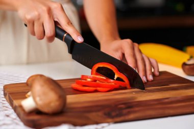 Bir kadın şef, taze mantar ve muzlarla çevrili ahşap kesme tahtasındaki kırmızı dolma biberi ustalıkla dilimliyor, canlı bir mutfak ortamındaki mutfak uzmanlığını vurguluyor.