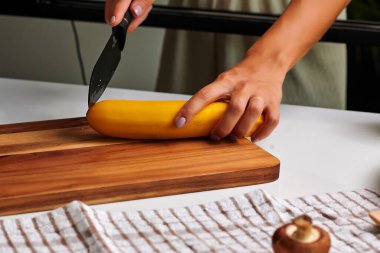 Bıçak tutan bir elin yakın çekimi, çağdaş bir mutfakta ahşap bir kesme tahtasının üzerinde taze sarı bir kabağı dilimlemek, canlı renkleri ve yemek hazırlama dokusunu vurgulamak,