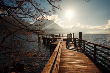 Manzaralı ahşap iskele, sakin su üzerine uzanır, dağlar ve ağaçlar tarafından çerçevelenir, sıcak güneş ışığıyla yıkanır, huzurlu bir doğal ortamı çağrıştırır.