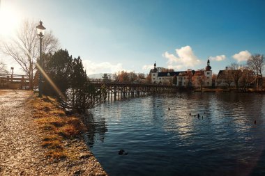 Ahşap bir köprüsü olan sakin bir sahil manzarası, bereketli ağaçlar ve güneş ışığıyla yıkanmış ve suya yansıyan tarihi binalar.