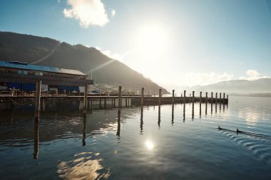 Sakin sulara uzanan ahşap bir iskelenin manzarası, arka planda dağlar ve güneş ışığı sakin bir atmosfer yaratıyor.
