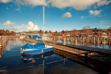 Mavi yelkenli durgun bir marinaya demir atmış, durgun suya yansıyor ve bulutlarla dolu güzel bir gökyüzü, huzurlu bir atmosfer yaratıyor.