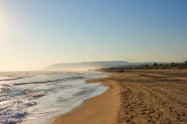 Sakin sahil sahnesinde, uzak tepeler ve açık gökyüzü ile altın kumlar üzerinde yumuşak dalgalar dinlenmek ve keyif almak için huzurlu bir atmosfer yaratır.