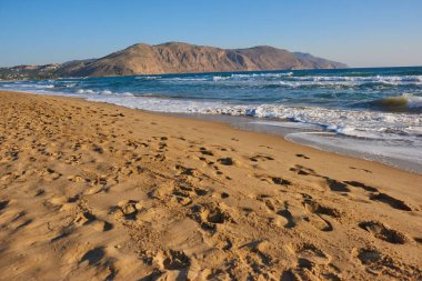 Kumsalda sakin dalgalara ve açık mavi gökyüzünün altındaki uzak tepelere giden ayak izleri var. Huzurlu bir kıyı sahnesini çağrıştırıyor.