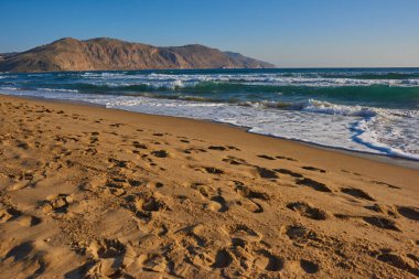 Yumuşak kum ve dalgaların olduğu sakin sahil manzarası. Ön planda parlak bir gökyüzünün altındaki dağlara giden ayak izleri.