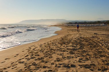 Huzurlu sahil sahnesinde kumlu kıyıya vuran hafif dalgalar var. Kıyı şeridinde gezinen yalnız bir figür, huzur ve rahatlığı çağrıştırıyor.