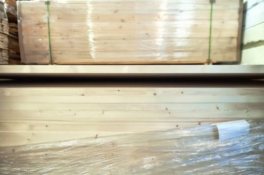 Plastiğe sarılı ahşap tahtalar, doğal ahşap dokular ve tahıllar sergileniyor, iyi aydınlatılmış bir depolama alanında inşaat malzemeleri vurgulanıyor.