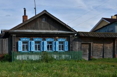 Kırsal ahşap evde mavi panjurlar ve yeşil bir çit var, canlı çimenlerin üzerine kurulmuş, geleneksel kırsal yaşamı ön plana çıkarıyor.