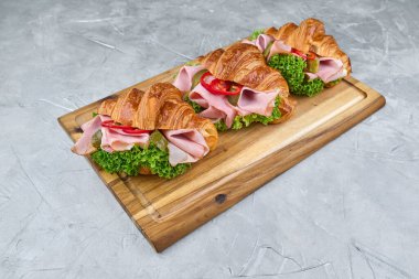 Jambon, kıtır marul ve kırmızı biber katmanlı kruvasan sandviçler, ahşap bir tahtada ustaca sergilenmiş, mutfak işçiliği sergileniyor.