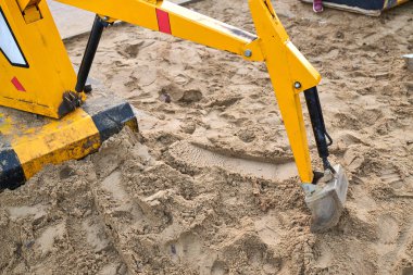 Kazı kolu aktif olarak kumlu araziyi kazıyor, inşaat malzemeleri ve aletleriyle ağır makinelerin gücünü vurguluyor.