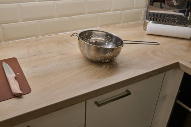 Paslanmaz çelik kevgir ahşap mutfak tezgahında oturur, mutfak gereçlerini ve mutfak aletlerini çağdaş bir yemek alanında renklendirir.