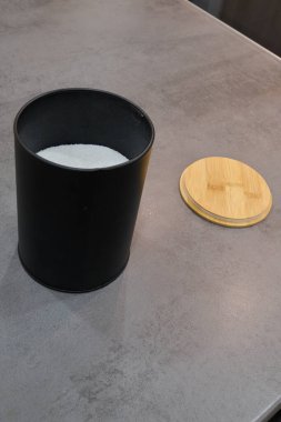 Siyah depolama kabı beyaz şeker içerir, bambu kapağı ile kaplıdır, şeffaf mutfak tezgahına kurulmuştur, çağdaş estetiği ve pratiği vurgular.