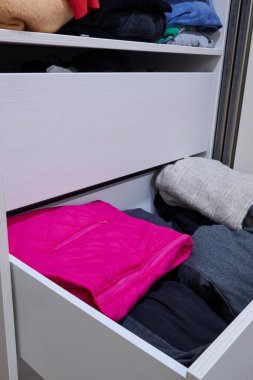Canlı pembe bir ceket belirgin bir şekilde çeşitli kıyafetlerle dolu beyaz bir çekmecede sergileniyor, düzenli bir düzen ve stili ön plana çıkarıyor.