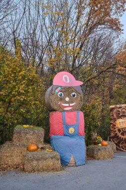 Gülen yüzlü, kırmızı şapkalı, mavi tulumlu, balkabaklarıyla ve sonbahar yapraklarıyla çevrili büyük bir saman balyası karakteri.