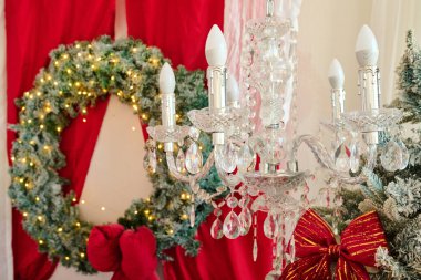 Işıklı süslü bir çelenk, kristal aksanlı bir avize ve kırmızı perdelerle süslenmiş bir bayram sahnesi.