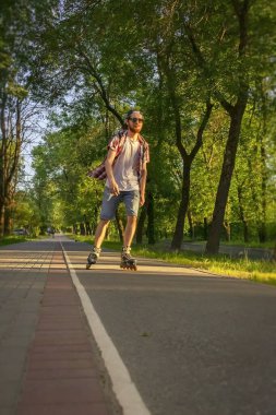 Ekose gömlekli, kot şortlu, güneş gözlüklü bir adam gün batımında şehirde paten kayıyor..