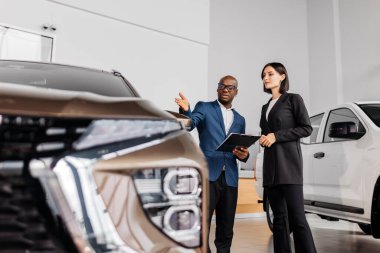 Erkek otomotiv satış temsilcisi modern bir galeride kadın müşteriyle araba özelliklerini tartışıyor, araç tasarımı ve profesyonel etkileşimi vurguluyor