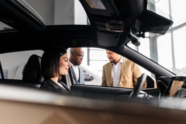 İki erkek satış temsilcisi, dinamik bir araba galerisi atmosferini ön plana çıkarırken, kadın müşteri şık bir araçta oturuyor ve gülümsüyor.