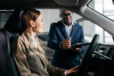 Arabadaki kadın takım elbiseli bir adamla etkileşime giriyor, çağdaş bir otomobil galerisinde araç özelliklerini tartışıyor, müşteri hizmetlerini ve nişan törenini gösteriyor.