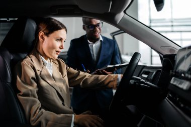 Bej takım elbiseli bir kadın profesyonel, modern bir aracın test sürüşünde erkek bir meslektaşıyla birlikte, otomotiv satış sürecinin altını çiziyor.