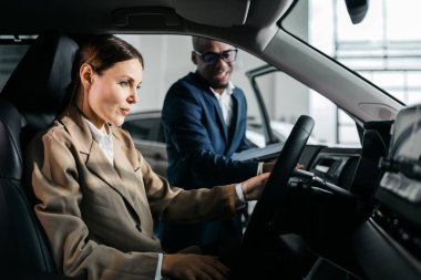 Bej ceketli kadın araç kontrolleriyle meşgul olurken, erkek satıcı da doğal ışıkla dolu çağdaş bir otomobil galerisinde ona yardım ediyor.