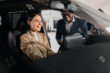 İş kadını, erkek meslektaşı ile aynı arabada oturuyor. Çağdaş bir otomotiv galerisinde araç özellikleri ve teknoloji sergiliyor. Profesyonel bir atmosfer yaratıyor.