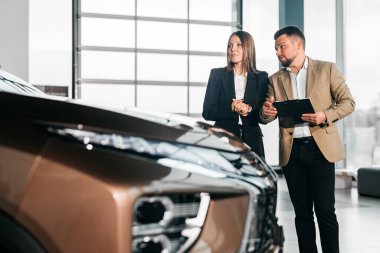 Çağdaş bir galeride kadın ve erkek lüks araba özellikleri hakkında konuşmakta, otomotiv tasarımı ve müşteri etkileşimini ön plana çıkarmaktadır.