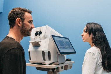 Erkek hasta, göz muayenesi sırasında kadın göz bakımı uzmanıyla etkileşime girer. Serin mavi bir ortamda gelişmiş teşhis ekipmanları kullanarak, sağlık alanındaki profesyonelliği vurgular.