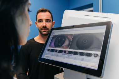 Kadın hasta göz muayenesiyle meşgul olurken, erkek teknisyen sağlık alanındaki yenilikleri ön plana çıkarırken, dijital görüntülere göz atmaktadır.