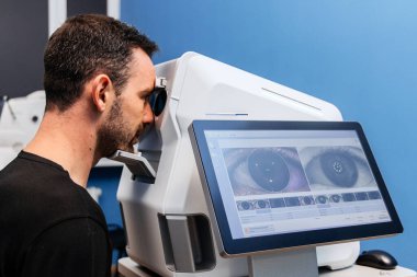 Erkek göz bakımı uzmanı dijital ekranda göz görüntülerini inceliyor, klinik ortamda gelişmiş ekipmanları kullanıyor, hassasiyet ve teknolojiyi vurguluyor.