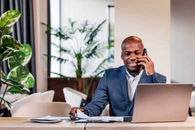 Profesyonel bir adam, şık ofis masasında dizüstü bilgisayar kullanırken telefonla konuşuyor. Bitkiler ve parlak pencerelerle canlı bir çalışma ortamı yaratıyor.