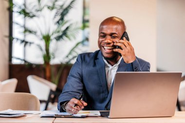 Mavi takım elbiseli gülümseyen erkek profesyonel, bir bilgisayar ve evrak işleriyle çağdaş bir ofis masasında, dinamik bir çalışma alanını yansıtan bir telefon görüşmesi yapıyor.