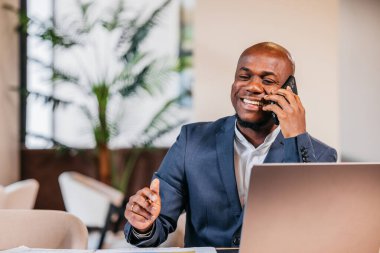 Erkek profesyonel, arka planda dizüstü bilgisayar ve yeşillik ile modern bir masada akıllı bir telefonla canlı bir çalışma alanı atmosferi yaratarak mutlu bir şekilde sohbet ediyor.