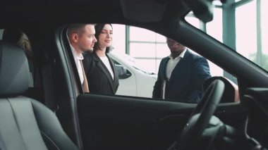 Bir galeride araba özellikleri, müşteri etkileşimleri ve bir araç satın almanın heyecanı hakkında sohbet eden çeşitli bireyler.