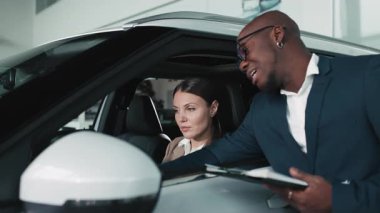 Bej ceketli kadın araç kontrolleriyle meşgul olurken, erkek satıcı da doğal ışıkla dolu çağdaş bir otomobil galerisinde ona yardım ediyor.