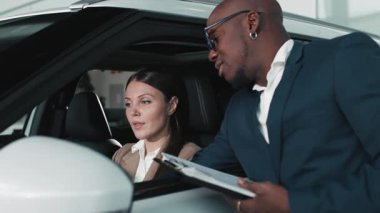 Bej ceketli kadın araç kontrolleriyle meşgul olurken, erkek satıcı da doğal ışıkla dolu çağdaş bir otomobil galerisinde ona yardım ediyor.