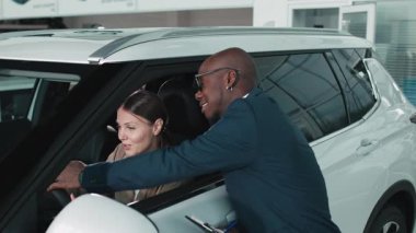 Bej ceketli kadın araç kontrolleriyle meşgul olurken, erkek satıcı da doğal ışıkla dolu çağdaş bir otomobil galerisinde ona yardım ediyor.