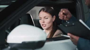 Bej ceketli kadın araç kontrolleriyle meşgul olurken, erkek satıcı da doğal ışıkla dolu çağdaş bir otomobil galerisinde ona yardım ediyor.