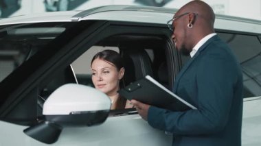 Bej ceketli kadın araç kontrolleriyle meşgul olurken, erkek satıcı da doğal ışıkla dolu çağdaş bir otomobil galerisinde ona yardım ediyor.