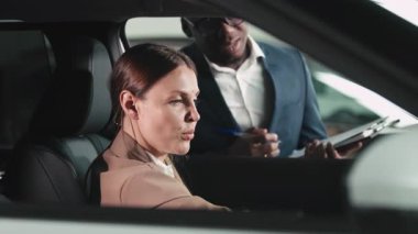 Bej ceketli kadın araç kontrolleriyle meşgul olurken, erkek satıcı da doğal ışıkla dolu çağdaş bir otomobil galerisinde ona yardım ediyor.