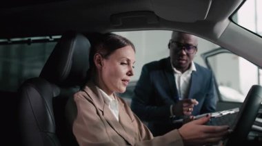 Bej ceketli kadın araç kontrolleriyle meşgul olurken, erkek satıcı da doğal ışıkla dolu çağdaş bir otomobil galerisinde ona yardım ediyor.