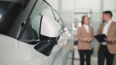 Pırıl pırıl beyaz bir arabanın arka planda iki profesyonelin bulunduğu parlak bir galeride araç özelliklerini tartışırken, müşteri etkileşimini ve servisi vurguluyor.