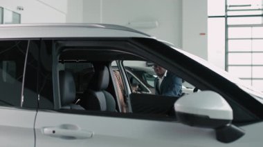 Takım elbiseli Afro-Amerikalı bir adam, bir galeride bir arabanın içinde beyaz bir kadınla takılıyor, özelliklerini ön plana çıkarıyor ve pozitif bir araba satın alma ortamı yaratıyor.