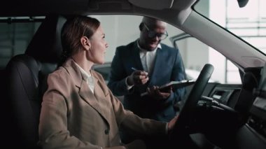 Takım elbiseli Afro-Amerikalı bir adam, bir galeride bir arabanın içinde beyaz bir kadınla takılıyor, özelliklerini ön plana çıkarıyor ve pozitif bir araba satın alma ortamı yaratıyor.