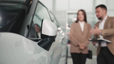 Pırıl pırıl beyaz bir arabanın arka planda iki profesyonelin bulunduğu parlak bir galeride araç özelliklerini tartışırken, müşteri etkileşimini ve servisi vurguluyor.