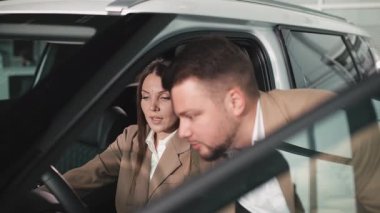 Nişanlı kadın ve bej takım elbiseli adam satış aracının içinde araba özelliklerini, müşteri etkileşimini ve araba satın alma deneyimini tartışıyor.