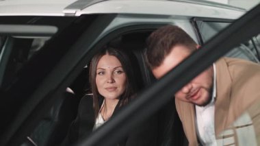 Nişanlı kadın ve bej takım elbiseli adam satış aracının içinde araba özelliklerini, müşteri etkileşimini ve araba satın alma deneyimini tartışıyor.