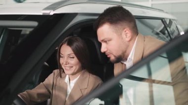 Nişanlı kadın ve bej takım elbiseli adam satış aracının içinde araba özelliklerini, müşteri etkileşimini ve araba satın alma deneyimini tartışıyor.